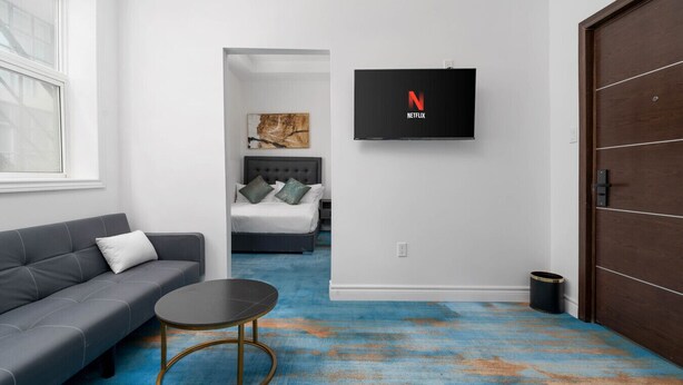 A hotel room with a grey sofa, a small round table, a bed with a tufted headboard, a blue carpet, a TV mounted on the wall, and a wooden door. - Executive Double Room ; Boutique Hotel• Trinity Bellwoods. Downtown Toronto (Toronto)