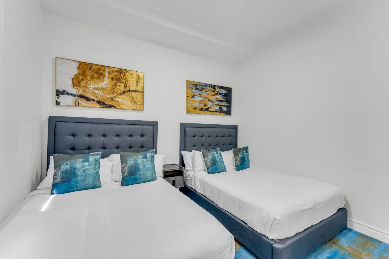 A hotel room with two beds, each with a grey headboard, white bedding, and blue accent pillows. There are two abstract paintings on the walls.