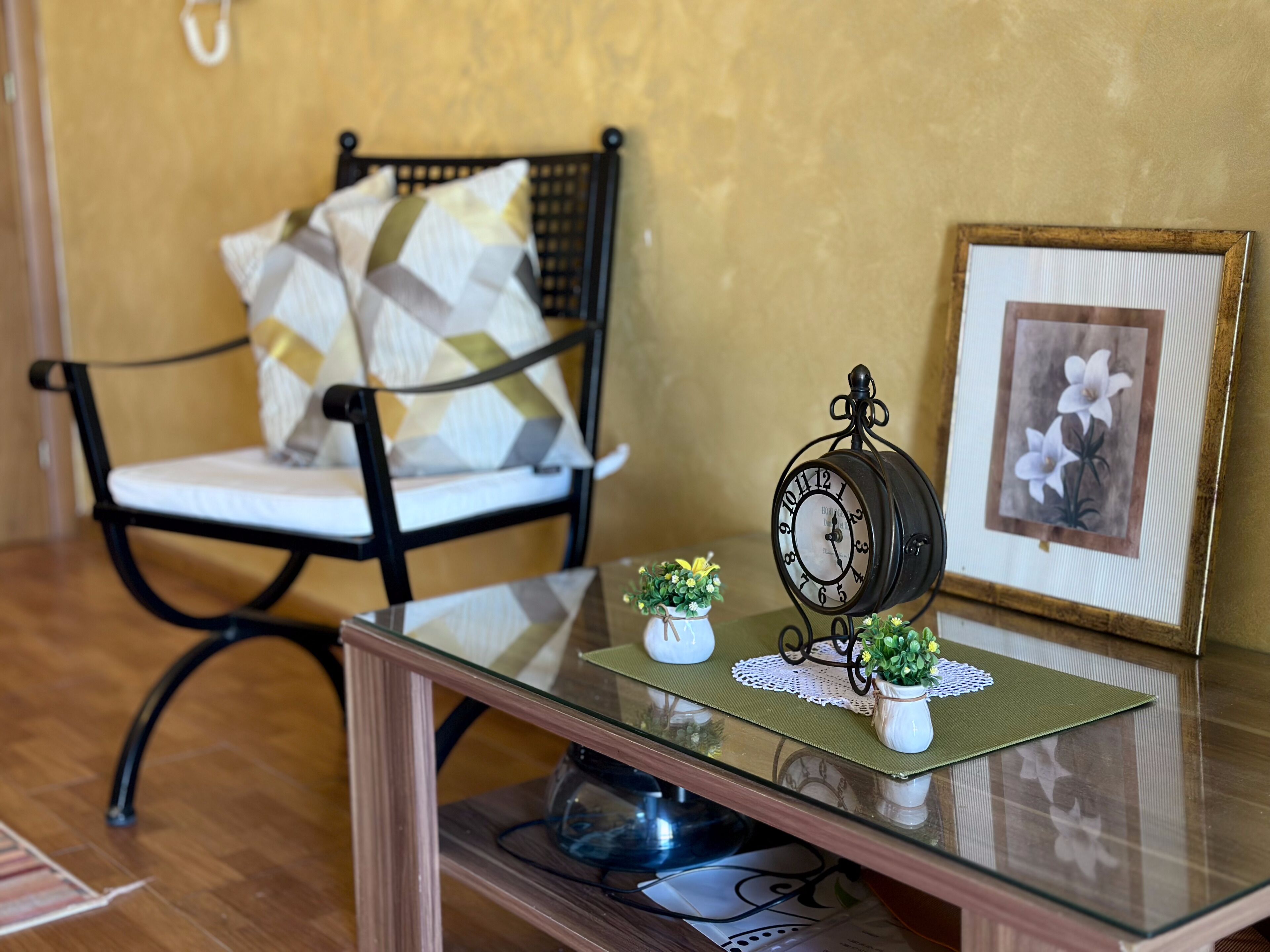 A chair with a patterned cushion, a small clock on a glass table, and a framed picture of flowers.