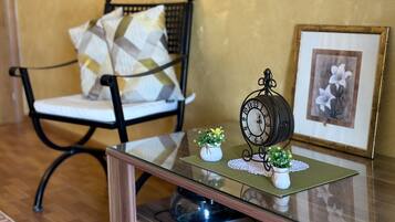 A chair with a patterned cushion, a small clock on a glass table, and a framed picture of flowers.