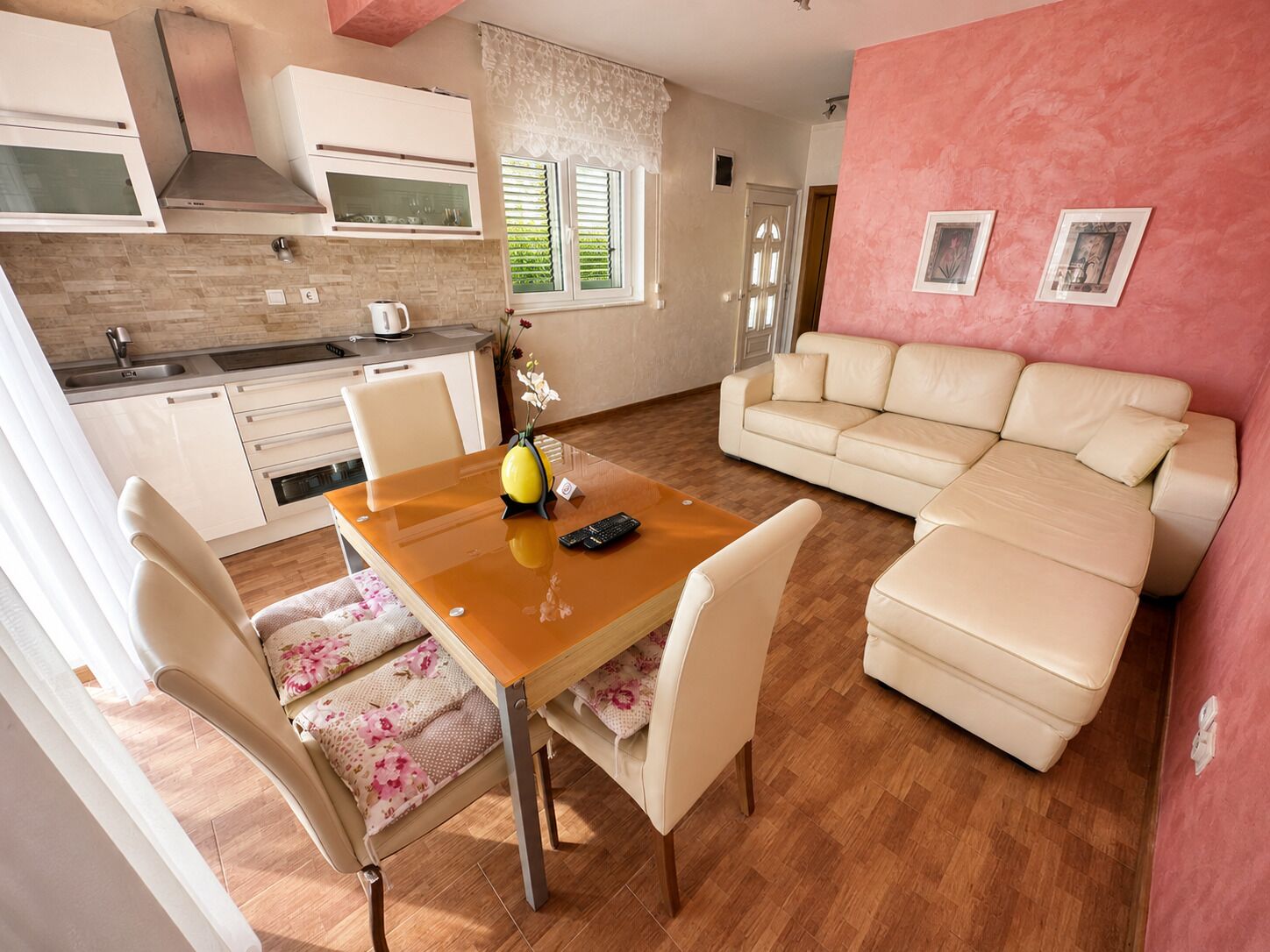 A modern kitchen with white cabinets, a wooden dining table with chairs, and a beige sofa in a living area with a pink wall.