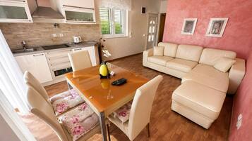 A modern kitchen with white cabinets, a wooden dining table with chairs, and a beige sofa in a living area with a pink wall.
