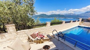 A poolside area with lounge chairs, a table with a vase of flowers, and a view of the sea and mountains. - Villa Mare (Krasici)