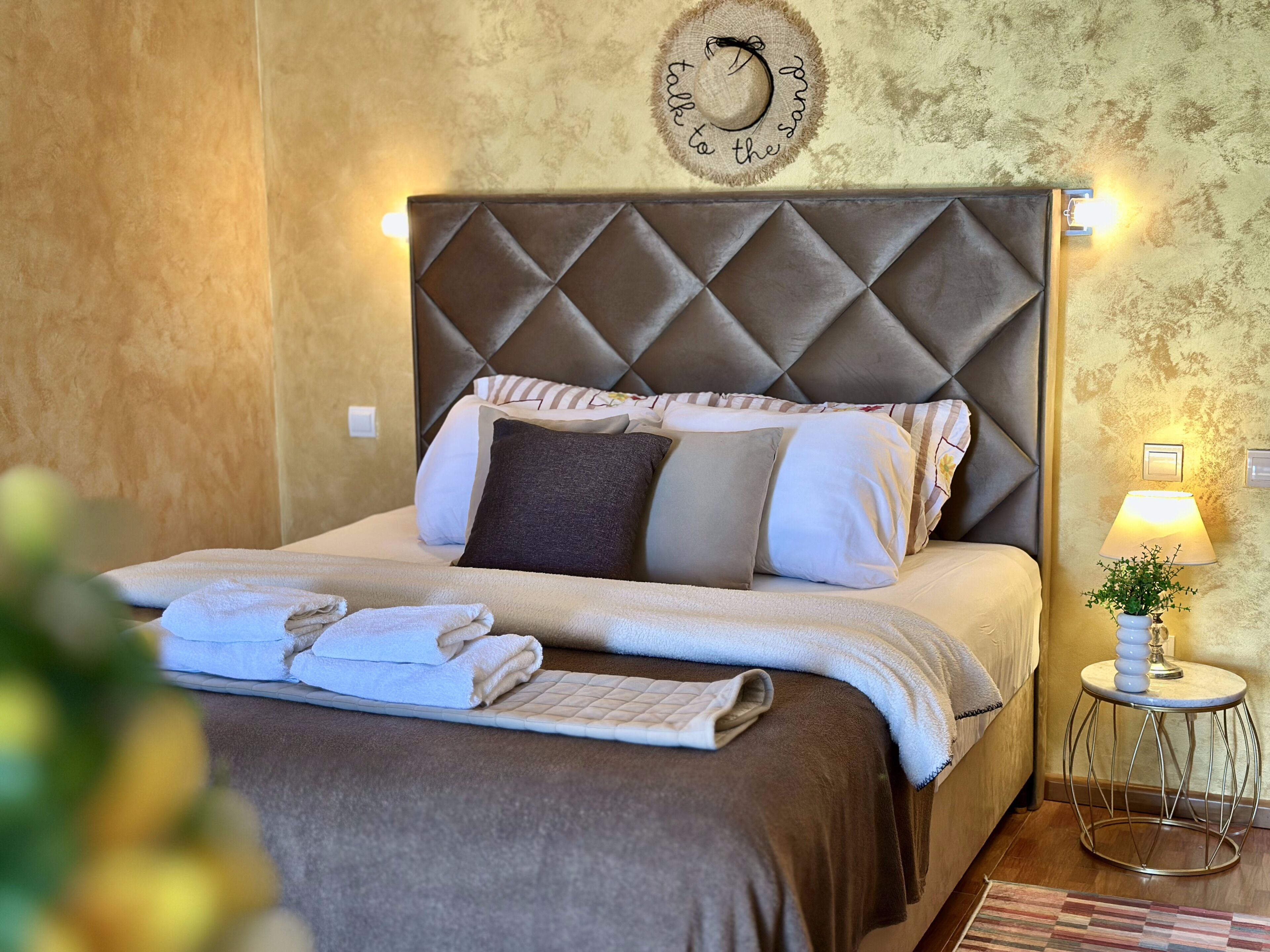 A neatly made bed with a quilted headboard, pillows, and a bedside table with a lamp and a small plant.