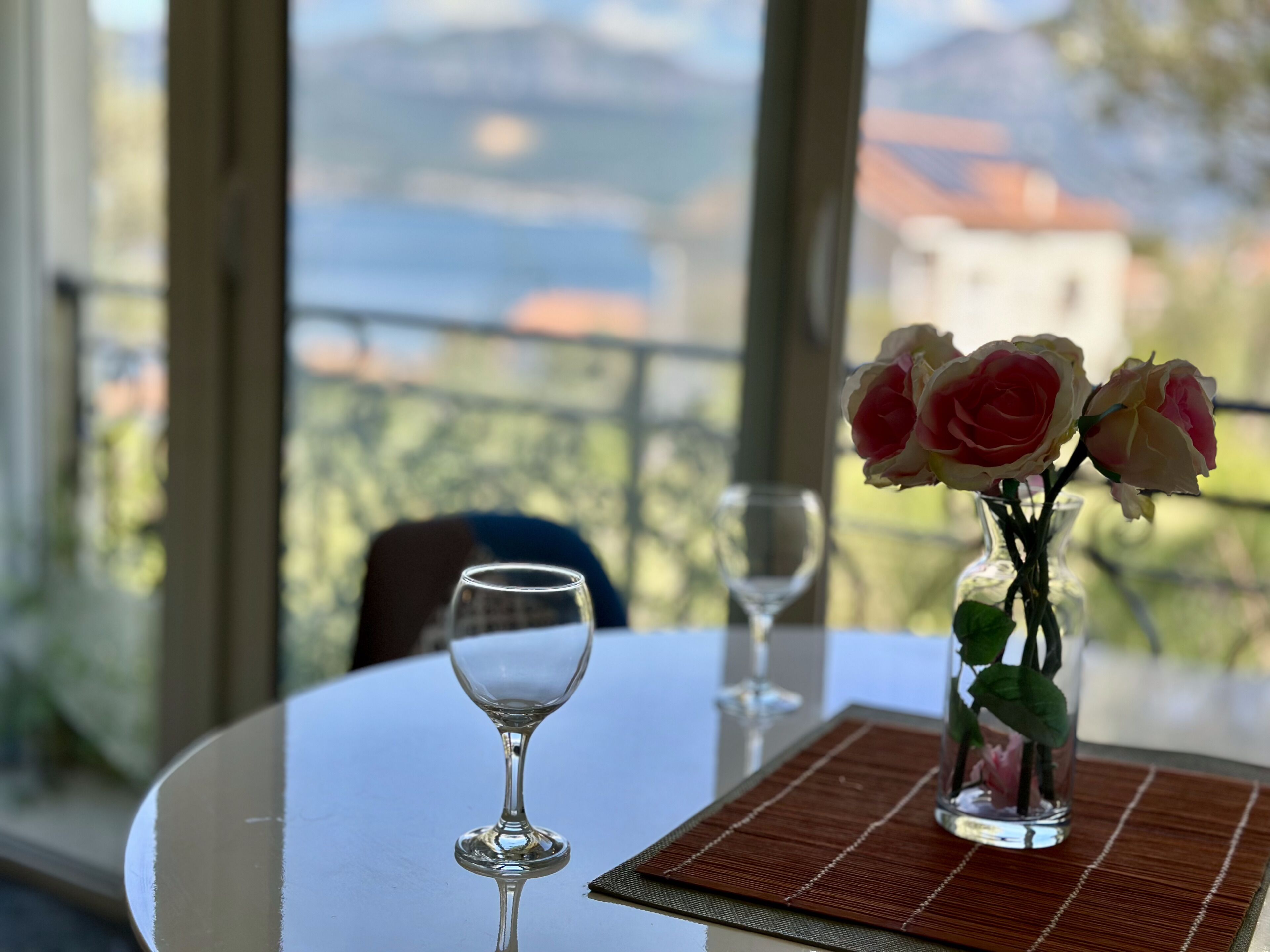 A table with a vase of flowers and two wine glasses, a clear view of a mountainous landscape through large windows.