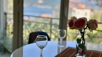 A table with a vase of flowers and two wine glasses, a clear view of a mountainous landscape through large windows.