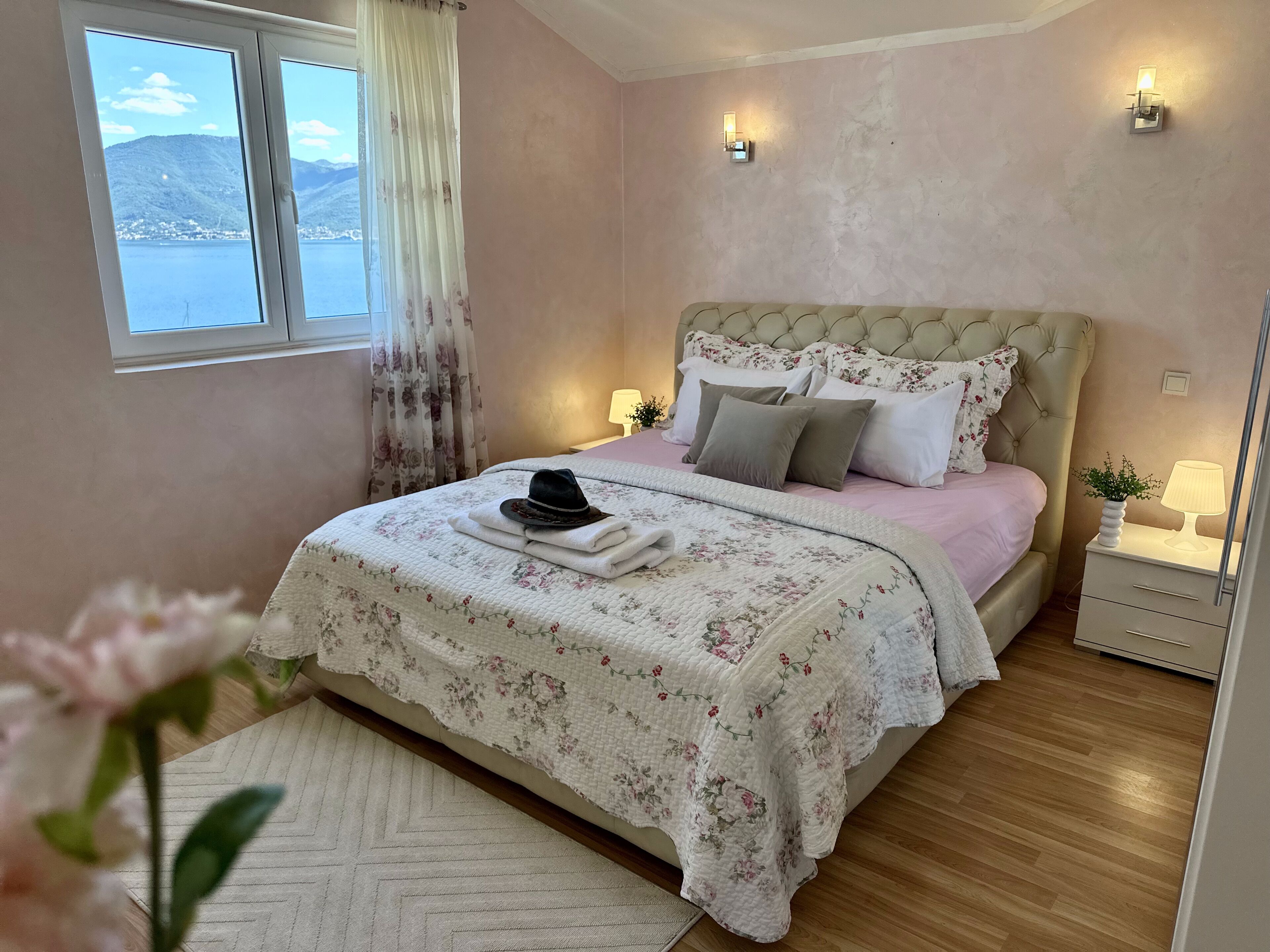 A bedroom with a bed, a nightstand, a lamp, a window with curtains, and a view of a body of water and mountains.