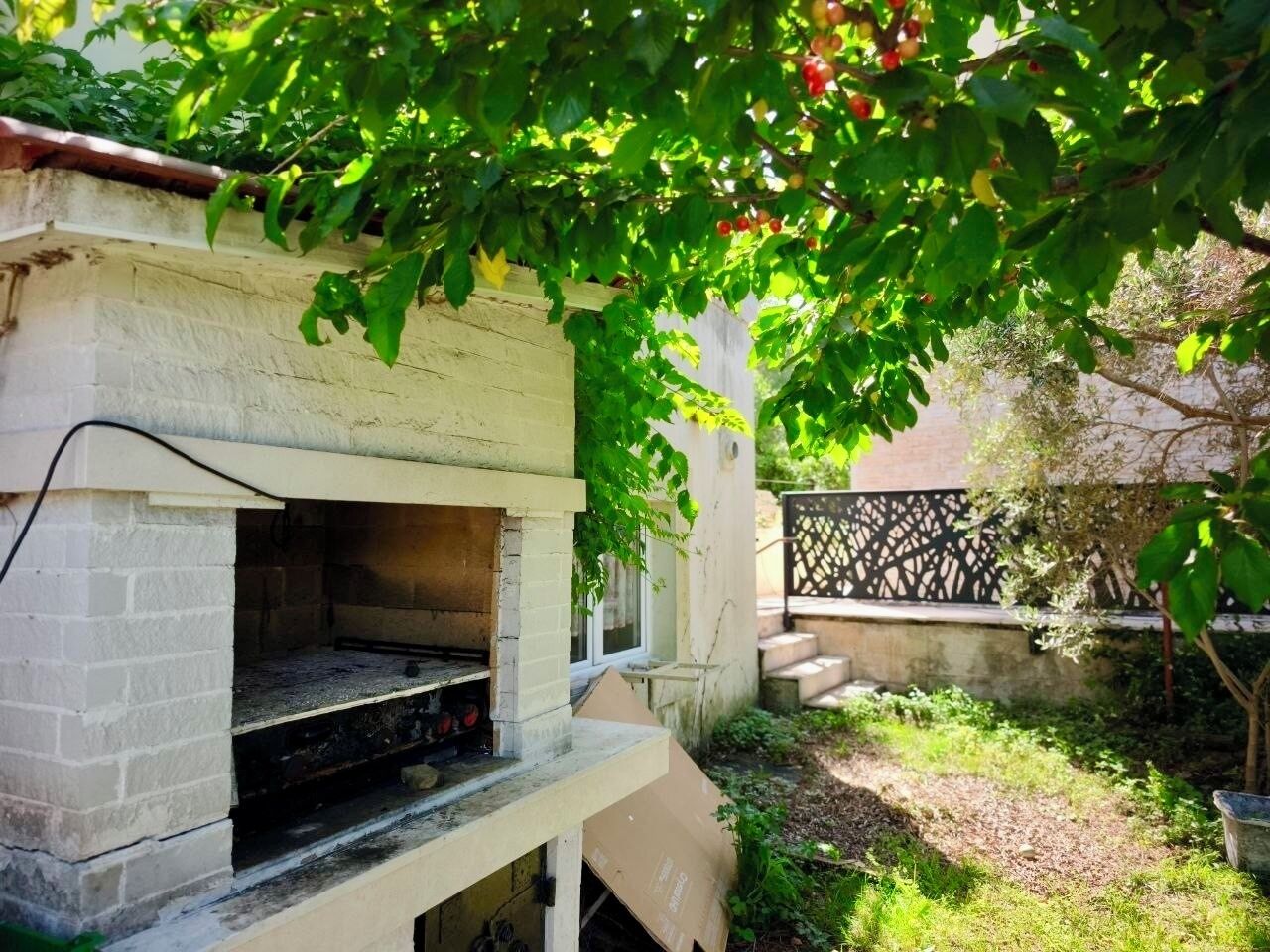 Un barbecue extérieur en briques, doté d’un four en briques, posé sur une base en béton et entouré de verdure et d’une clôture.