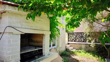 Un barbecue extérieur en briques, doté d’un four en briques, posé sur une base en béton et entouré de verdure et d’une clôture.