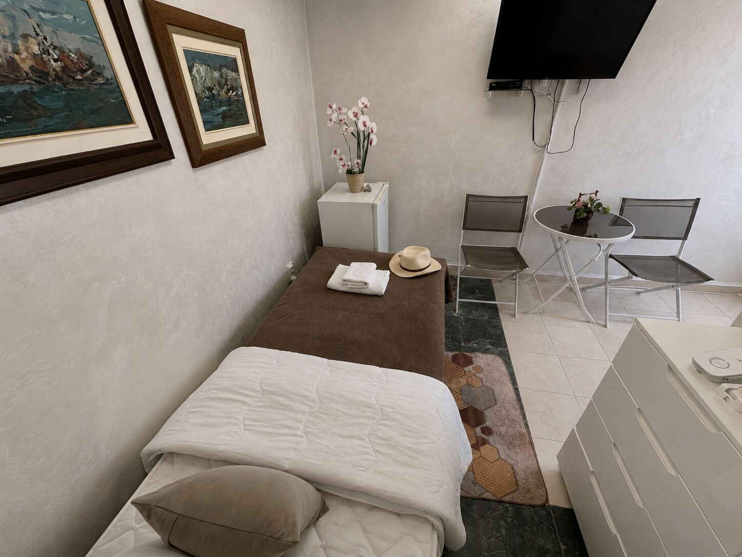 A single bed with a brown blanket, a white pillow, and a beige hat on it. There is a small white table with a vase of flowers and a white chair next to it. A television is mounted on the wall above the table.