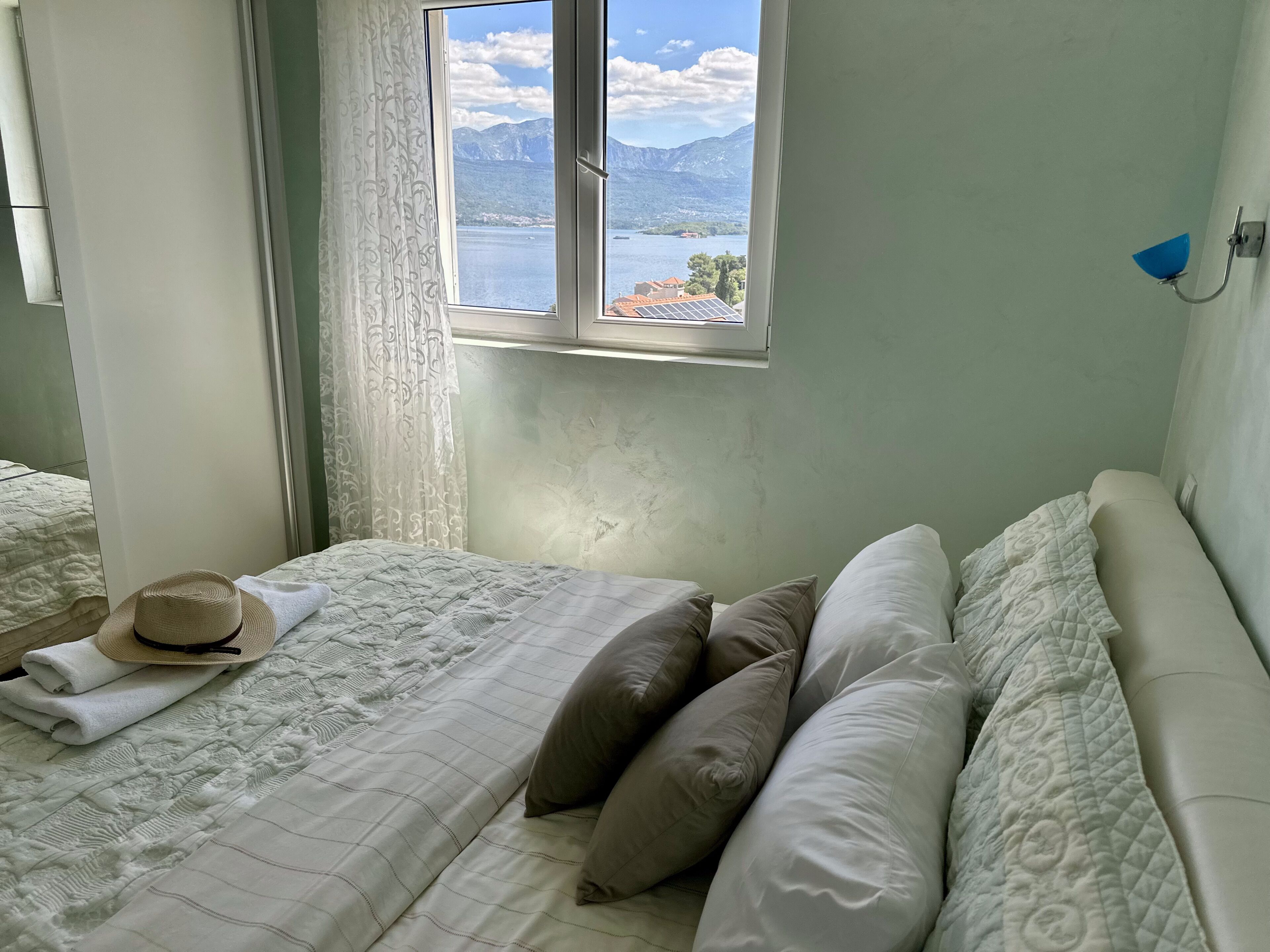 A bedroom with a bed, pillows, a hat, and a window with a view of mountains and a lake.