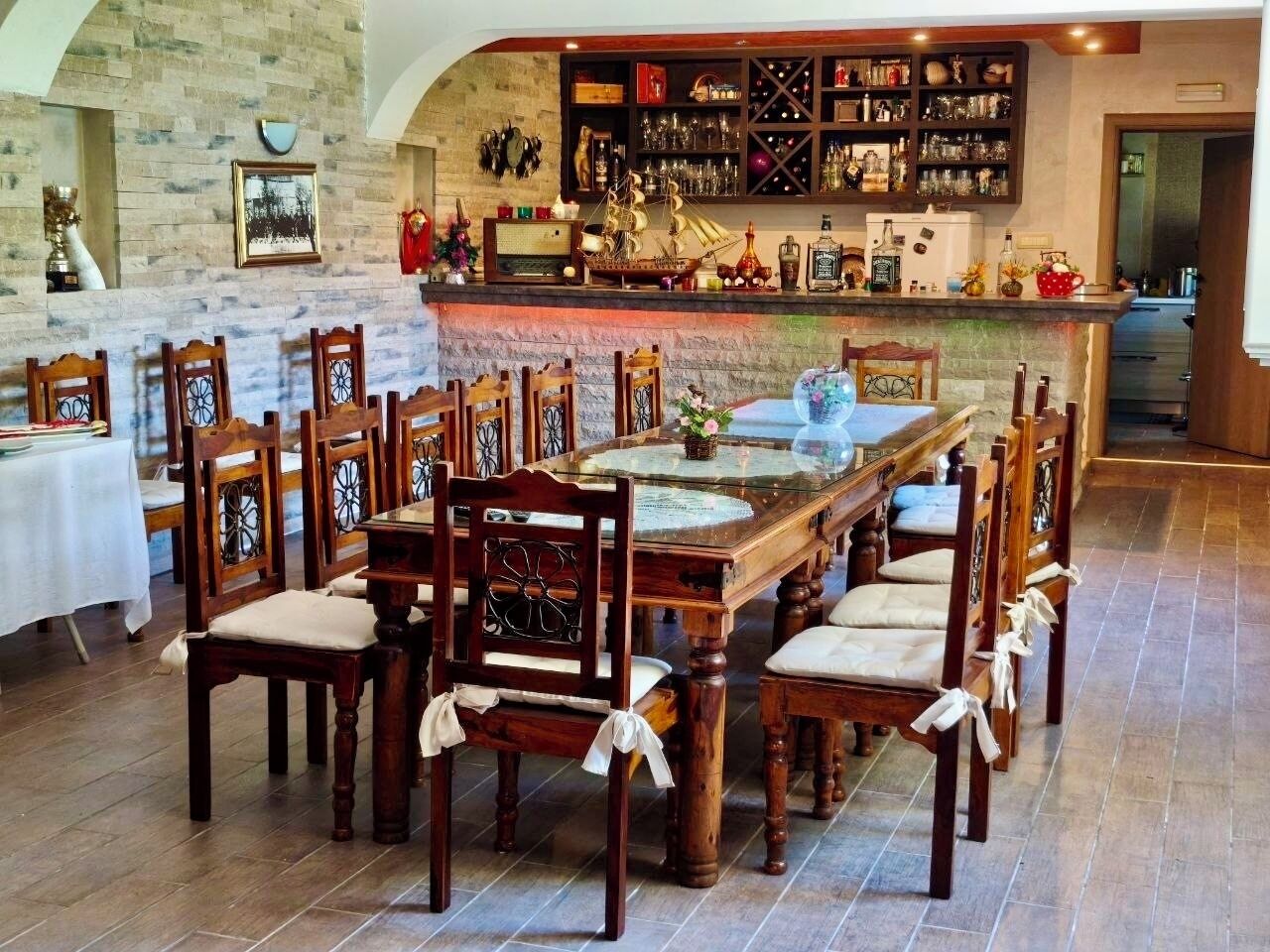 A dining area with a long wooden table surrounded by chairs, a bar with shelves of bottles and glasses, and a stone wall with framed pictures.