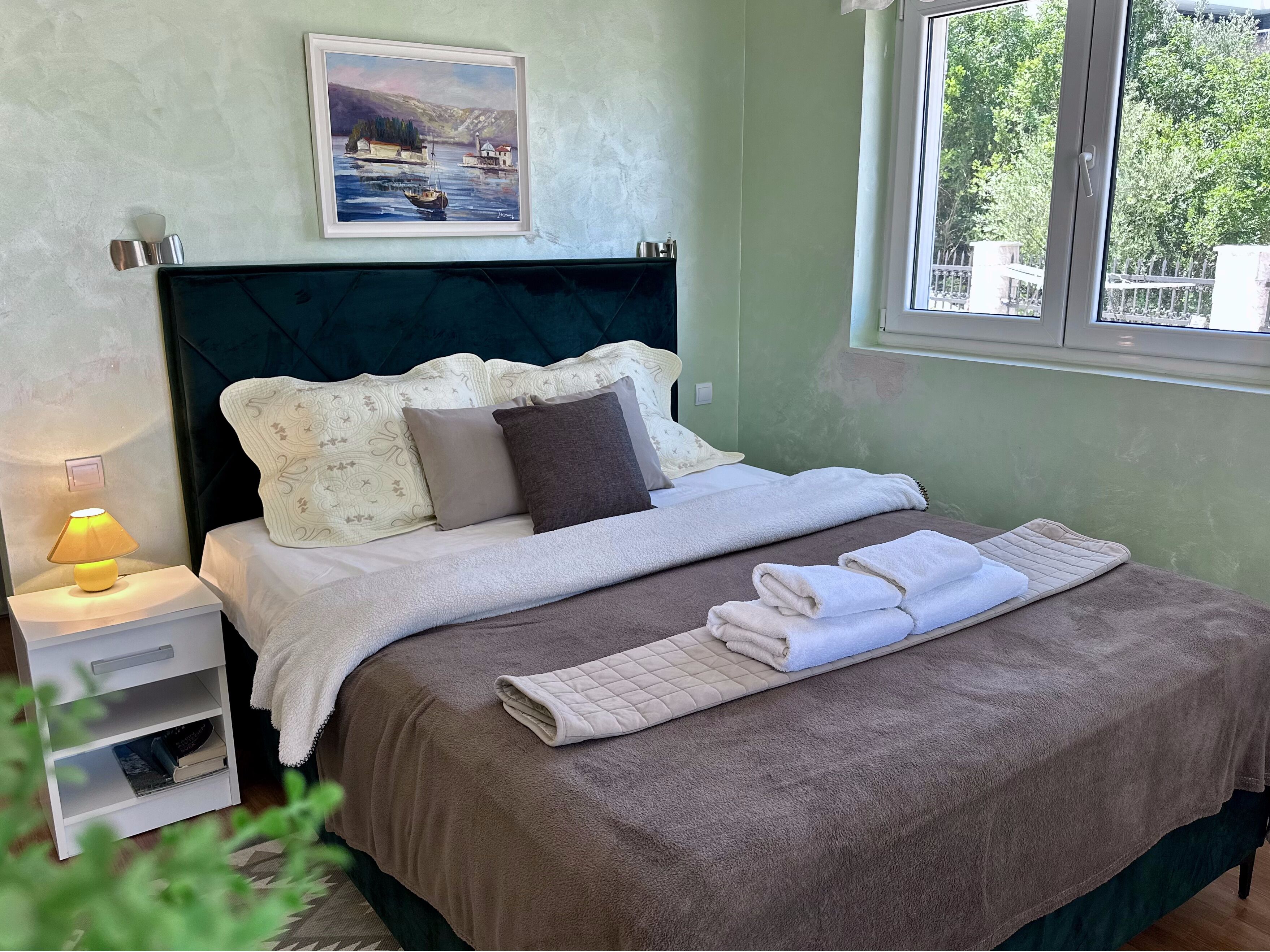 Un lit bien fait, recouvert d’une couverture marron, avec des oreillers et une serviette pliée. On y trouve une table de chevet avec une lampe, un tableau encadré au-dessus du lit et une fenêtre donnant sur un paysage verdoyant.