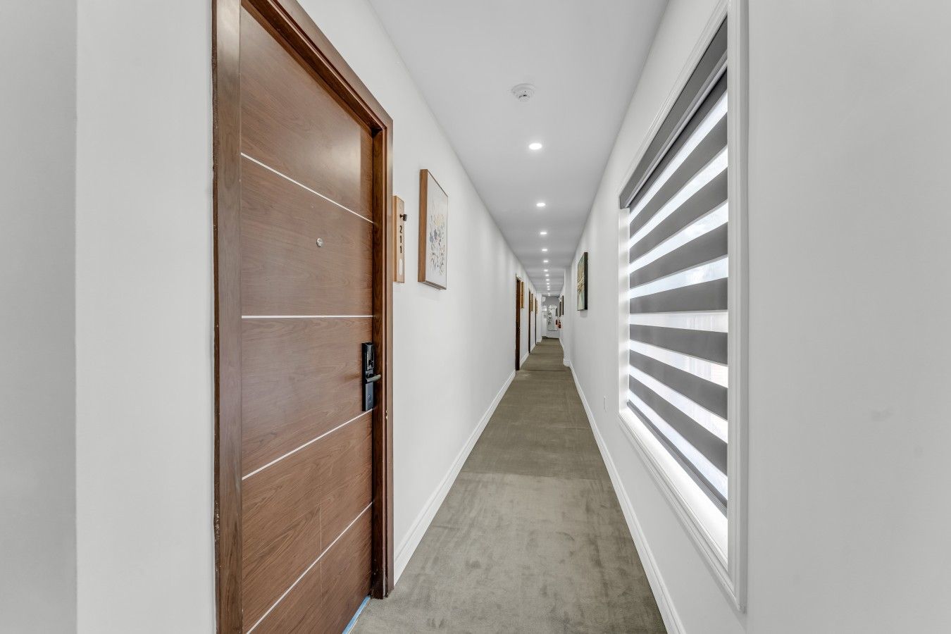 A corridor with a wooden door, a window with blinds, and a ceiling with recessed lighting.