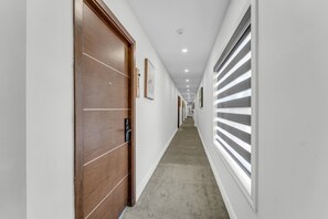A corridor with a wooden door, a window with blinds, and a ceiling with recessed lighting.