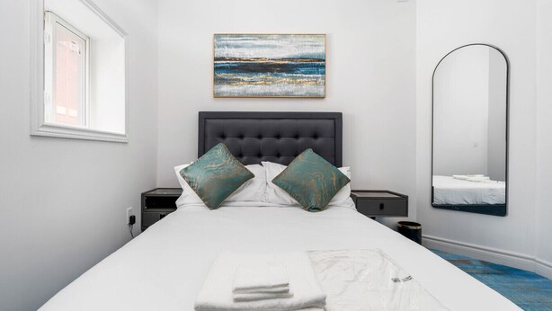 A neatly made bed with pillows, a bedside table, a nightstand, a painting, and a mirror.