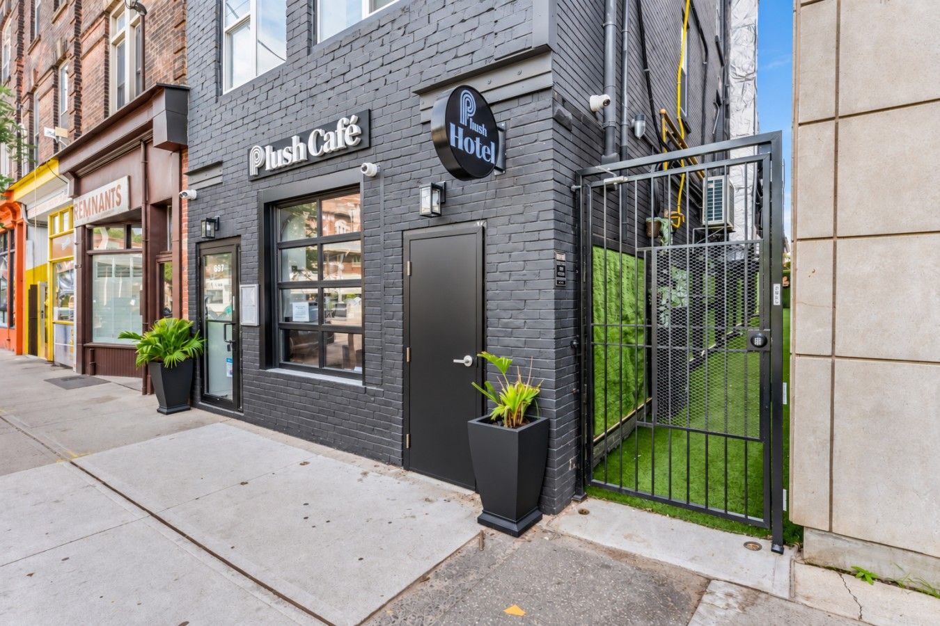 A building with a black exterior, a 'Push Café' sign, and a 'Blush Hotel' sign. There are two potted plants in front of the entrance.