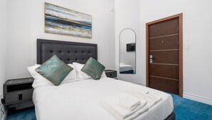 A neatly made bed with white linens and teal pillows, a bedside table with a lamp, a wall-mounted artwork, and a wooden door.