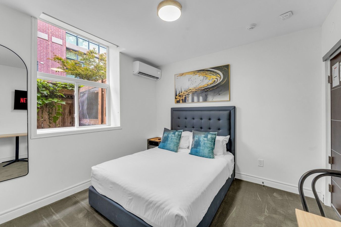 Un dormitorio con cama, ventana, aire acondicionado, un cuadro y una puerta.