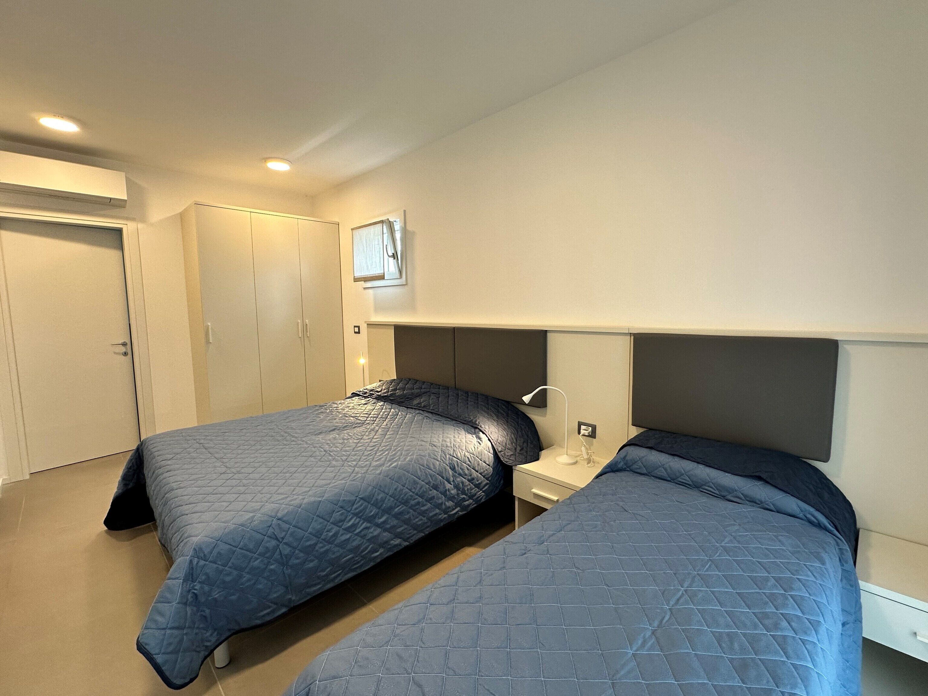 Quarto com duas camas, guarda-roupa, mesa de cabeceira e luminária no teto.
