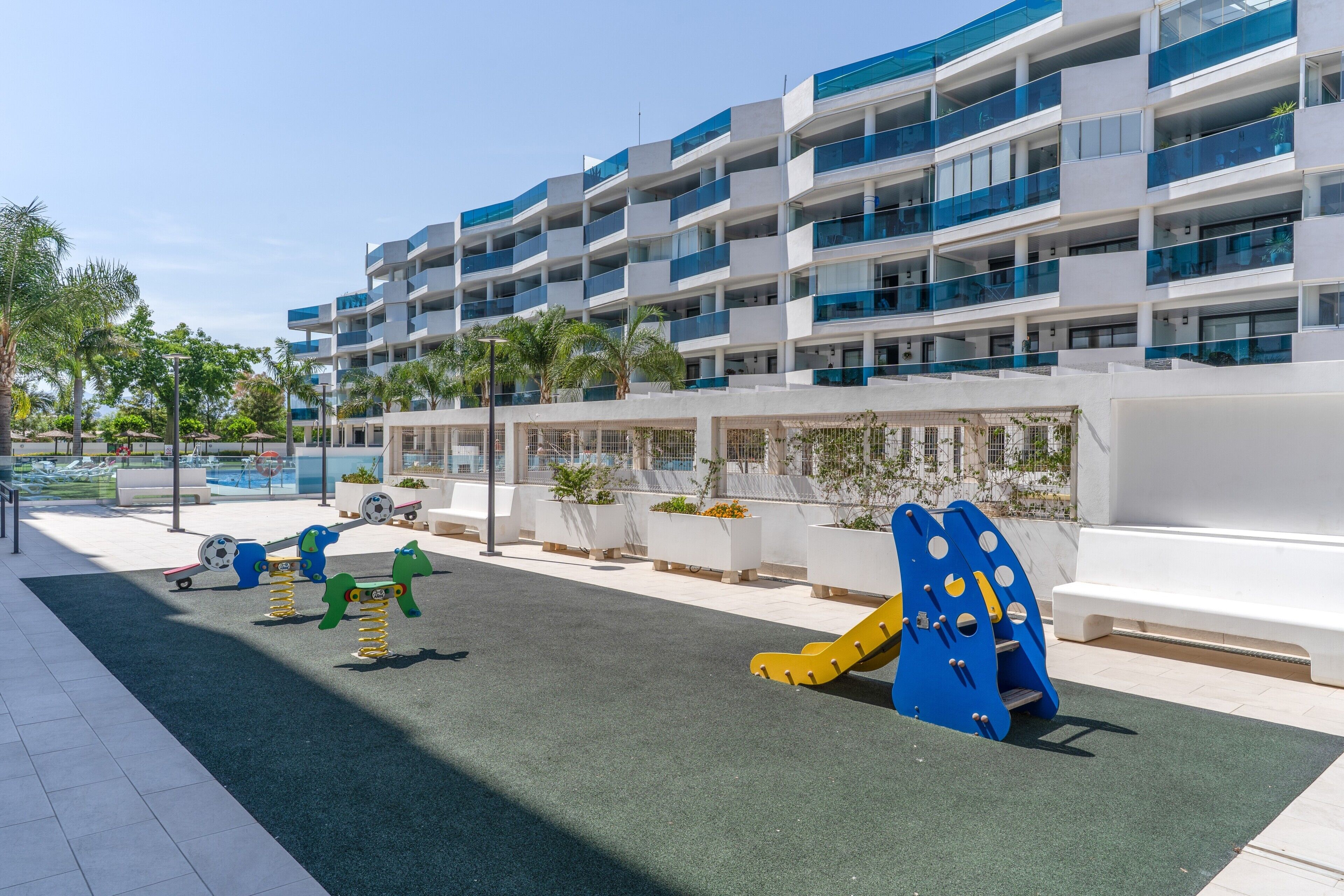 Un parque infantil con columpios y estructuras para escalar, una piscina y un edificio moderno con balcones.