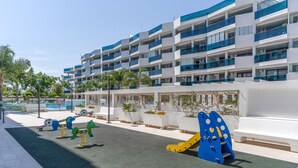 Ein Spielplatz mit Schaukeln und Klettergerüsten, ein Schwimmbad und ein modernes Gebäude mit Balkonen.