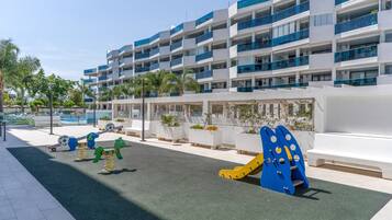 Un parque infantil con columpios y estructuras para escalar, una piscina y un edificio moderno con balcones.