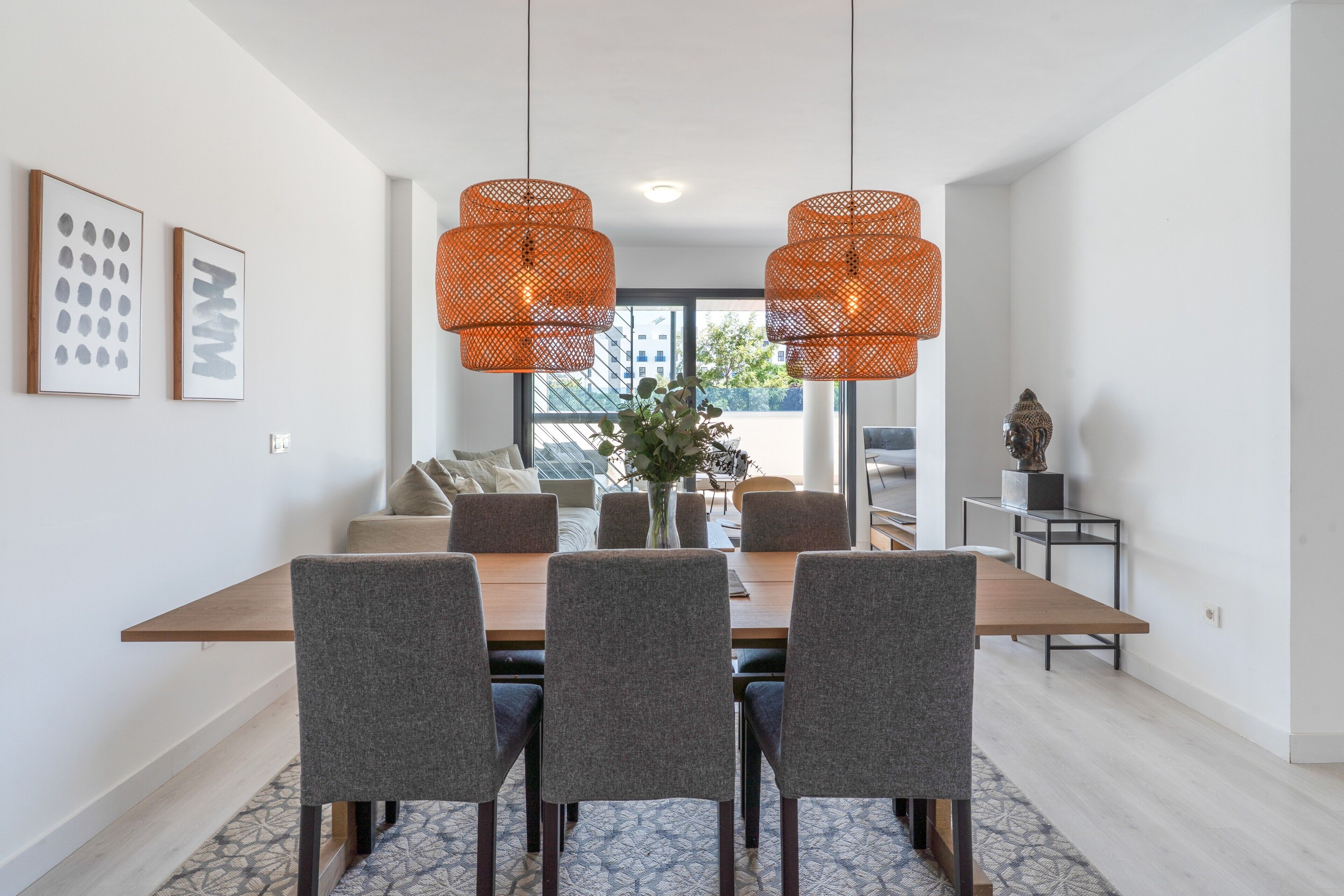Un comedor con mesa de madera, sillas tapizadas en gris y lámparas colgantes de fibras anaranjadas.