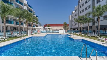 Una piscina con sillones de descanso y palmeras, rodeada de edificios modernos.