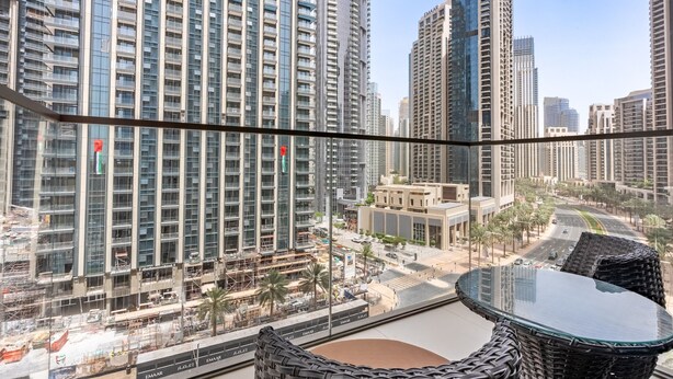 A high-rise building with a glass facade, a balcony with wicker furniture, and a view of a cityscape with palm trees and a road. - Breathtaking Skyscraper Views 3BR, Just a Stone Throw from Burj Khalifa, Dubai Mall (Dubai)