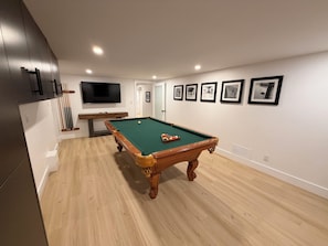 A pool table in a room with wooden flooring, a television mounted on the wall, and framed pictures on the wall.