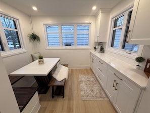 A modern kitchen with white cabinets, a wooden bench, a dining table, and a window with blinds.