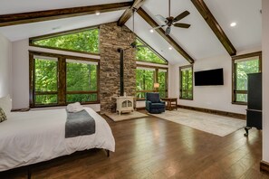 Un dormitorio amplio con una cama grande, una pared de piedra, una chimenea, un ventilador de techo y ventanas grandes.