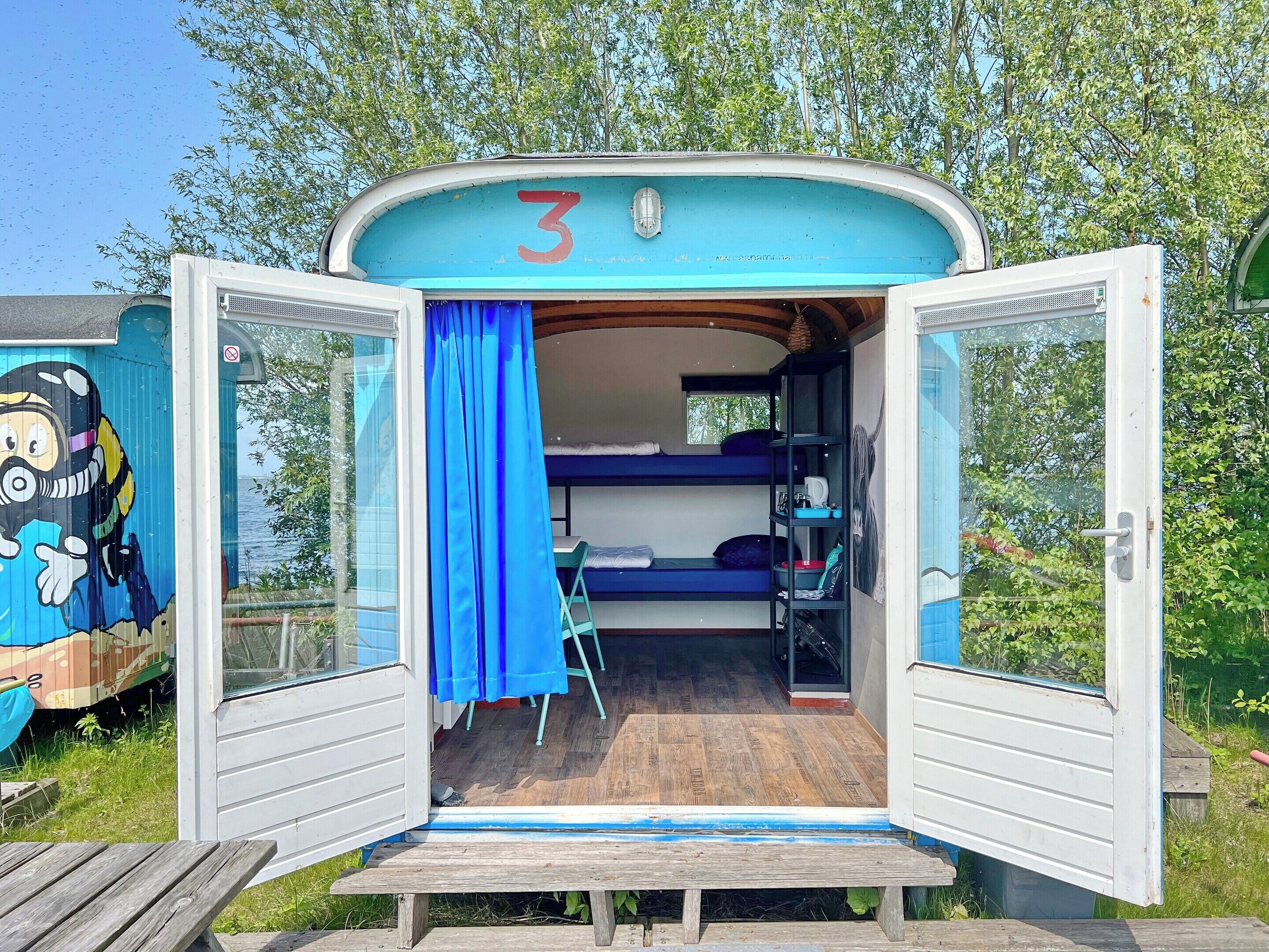 Un petit camping-car ouvert, avec le chiffre 3 peint sur la façade, un lit superposé à l’intérieur et une terrasse en bois à l’extérieur.