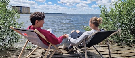 Two people sitting on wooden deck chairs facing a body of water with a building in the background.