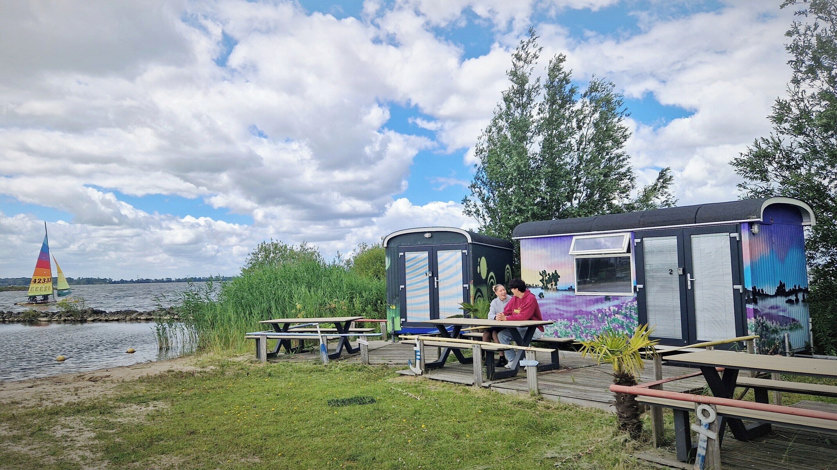 A lakeside campsite with mobile homes, picnic tables, and a sailboat.