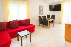 Ein Wohnzimmer mit einem roten Sofa, einem kleinen Tisch, einem Esstisch mit Stühlen, einem Fernseher an der Wand und einem Bild an der Wand.