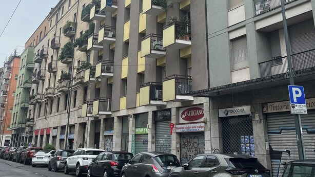 A multi-story residential building with balconies, parked cars, and storefronts. - Three-room apartment de Angeli area (Milano)