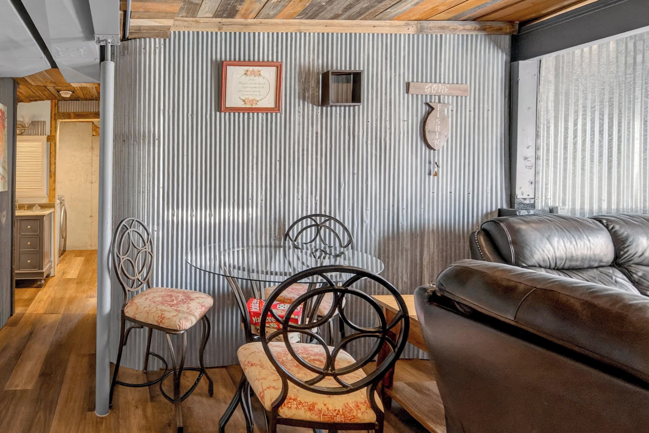A room with a wooden ceiling, a metal-framed table with chairs, a leather sofa, and a corrugated metal wall with a framed certificate and a sign.