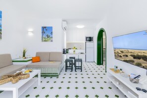 Un espacio habitable compacto con kitchenette, un sofá, comedor y un televisor que muestra un paisaje desértico.