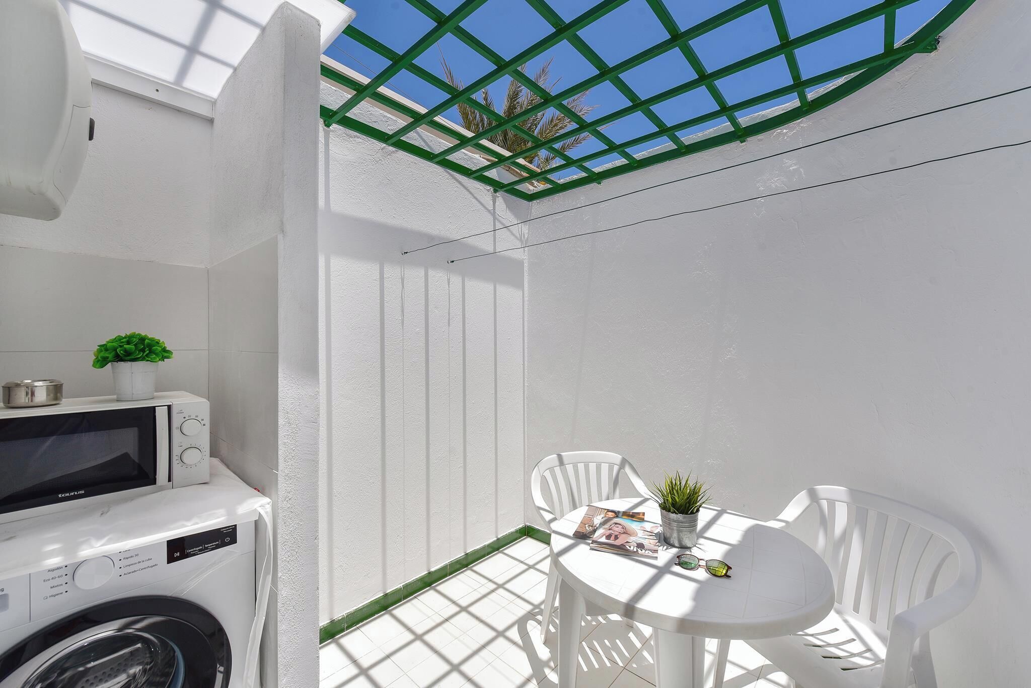 A compact, white-walled room with a washing machine, a small table with chairs, and a green-framed window.