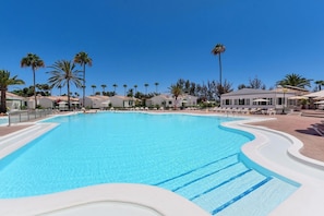 Ein großer Pool mit klarem, blauem Wasser, umgeben von Palmen und weißen Gebäuden mit grauen Dächern.