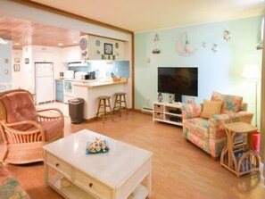 A living room with a sofa, a chair, a coffee table, a TV, and a kitchen area in the background. - Direct ocean front 1 BR and Den, Pool, Parking, WiFi Beautifully Decorated (Ocean City)