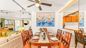 Un comedor con una mesa de madera dispuesta para cuatro personas, un ventilador de techo y un cuadro de veleros.