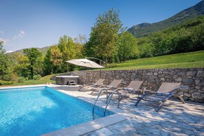 Ein Swimmingpool mit Liegestühlen und einer Steinmauer, umgeben von Grünflächen und Bergen.