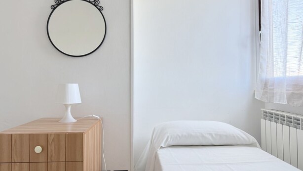 Ein Schlafzimmer mit einem Nachttisch aus Holz, einer weißen Lampe, einem runden Spiegel und einem Bett mit weißer Bettwäsche.