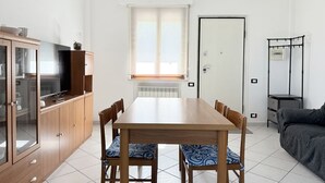 Ein gemütlicher Wohnraum mit Esstisch, Stühlen, einem Fernseher auf einem Schrank, einer Heizung, einer Tür und einem kleinen Beistelltisch.