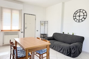 A room with a wooden dining table, a grey sofa, a wall clock, and a radiator.