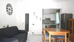 Ein Wohnzimmer mit einer Couch, einem Esstisch mit Stühlen, einer Küchenzeile mit Schränken und Geräten sowie einer Wanduhr.
