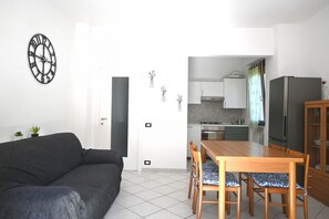 A living room with a sofa, a dining table with chairs, a kitchen area with cabinets and appliances, and a wall-mounted clock.